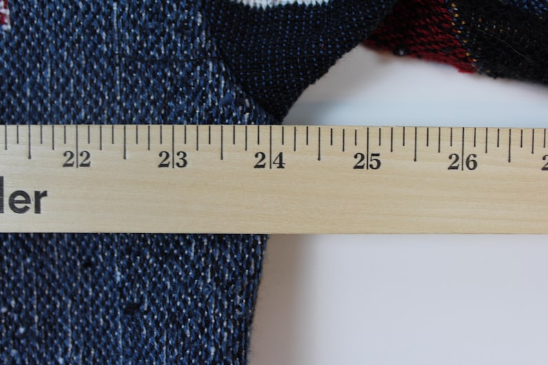 Op de afbeelding: Een blauw en zwart gebreid kledingstuk met een geweven textuurpatroon. Een houten liniaal is horizontaal over de stof geplaatst, waarbij de maatstreepjes van 55 tot 69 centimeter duidelijk zichtbaar zijn en de breedte van het item aangeven.