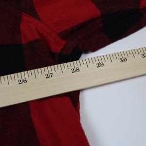 May include: Close-up of a red and black plaid flannel shirt. A wooden ruler is placed diagonally across the shirt, with measurements in inches. The shirt's fabric is visible, showing the texture and pattern.