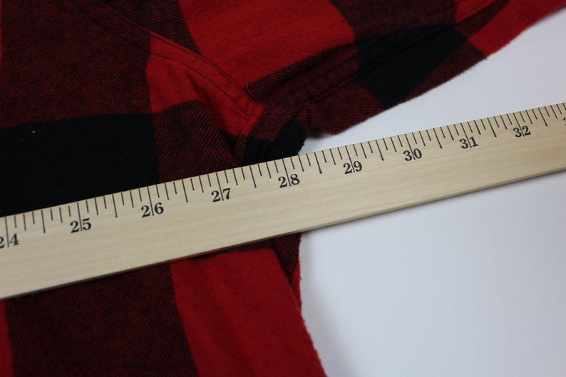 May include: Close-up of a red and black plaid flannel shirt. A wooden ruler is placed diagonally across the shirt, with measurements in inches. The shirt's fabric is visible, showing the texture and pattern.