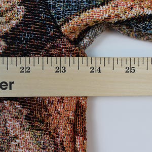 May include: A woven fabric garment featuring a multi-colored floral pattern in shades of orange, black, and blue. A wooden ruler is positioned horizontally across the fabric, indicating a width measurement of approximately 5.5 inches.