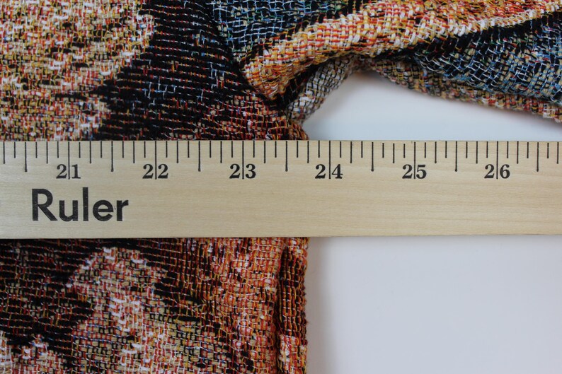 May include: A woven fabric garment featuring a multi-colored floral pattern in shades of orange, black, and blue. A wooden ruler is positioned horizontally across the fabric, indicating a width measurement of approximately 5.5 inches.
