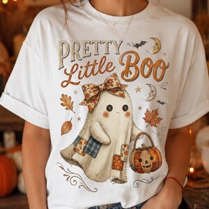 Op de afbeelding: Een wit T-shirt met korte mouwen en een afbeelding van een geest met een strik met patroon die een pompoenemmer vasthoudt. De tekst luidt Pretty Little Boo, vergezeld van illustraties van herfstbladeren, sterren en maansikkels.