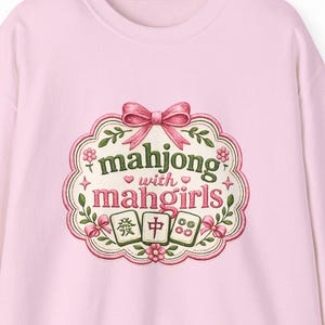 Puede incluir: Una sudadera rosa claro con un diseño floral en la manga izquierda. La parte delantera presenta un emblema decorativo con el texto "mahjong with mahgirls" en una fuente caprichosa, un lazo rosa y detalles florales. El emblema también incluye caracteres chinos.