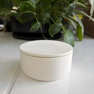May include: A circular white container with a flat, matching lid. The object has a smooth, matte finish and a simple, cylindrical shape. It is positioned as a standalone item, showcasing a clean, solid white colour throughout its entire exterior surface.