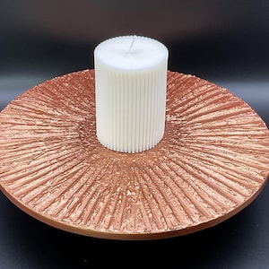 Op de afbeelding: Een witte, geribbelde pilaar kaars staat op een koperkleurige, getextureerde decoratieve schaal. De schaal heeft een radiaal patroon dat uit het midden komt. De kaars en schaal staan tegen een donkere achtergrond, wat een opvallend contrast creëert.