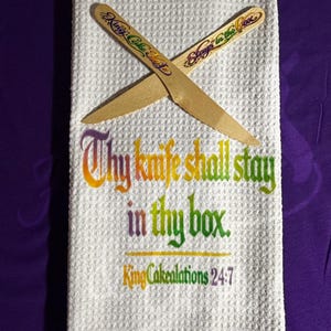 May include: A white waffle-weave kitchen towel with the text "Thy knife shall stay in thy box" in colorful lettering. Two wooden knives are crossed on top. The towel also has the text "King Cakealations 24:7".