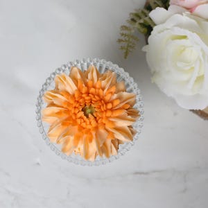 以下が含まれることがあります： オレンジ色の花の模様が入った透明なガラスボウルの俯瞰図。花は詳細な花びらの配置とテクスチャード加工された中心部を持っています。ボウルにはビーズの縁があります。背景には人工の白いバラがあります。