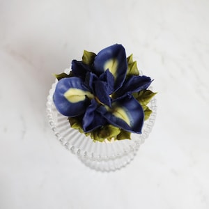 以下が含まれることがあります： バタークリームの花のデザインのカップケーキ。花は、黄色の中心と緑の葉を持つ濃い青色の花びらを持っています。カップケーキは、スカラップエッジの透明なガラスの台座にあります。