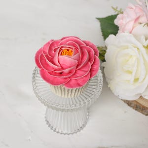 以下が含まれることがあります： バラの形をしたピンク色のカップケーキで、黄色の中心があり、透明なガラスの台座の上に置かれています。 カップケーキが焦点で、背景には白とピンクの花があります。