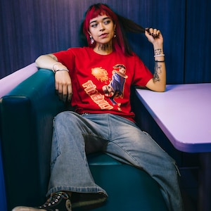 May include: A red t-shirt with a graphic and the text "Firebird". The person is wearing blue jeans and black platform trainers. The person is sitting in a teal booth. The image has a retro vibe.