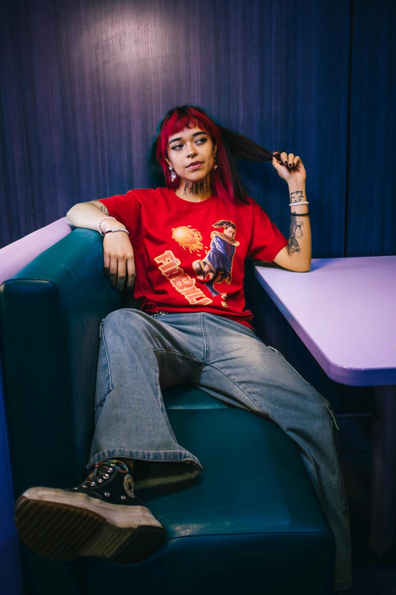 May include: A red t-shirt with a graphic and the text "Firebird". The person is wearing blue jeans and black platform trainers. The person is sitting in a teal booth. The image has a retro vibe.