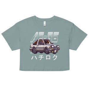 May include: A cropped, seafoam green t-shirt featuring a white and silver car graphic with Japanese text. The car is a stylized illustration of a classic sports car. The text above the car is in Japanese characters, and the text below the car reads "ハチロク".