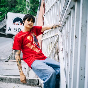 May include: A person wearing a red t-shirt with a graphic and the word "FIREBALL" in orange and white. The person is wearing blue jeans and has tattoos on their arms. They are leaning against a white metal fence.