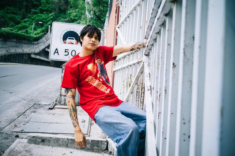 May include: A person wearing a red t-shirt with a graphic and the word "FIREBALL" in orange and white. The person is wearing blue jeans and has tattoos on their arms. They are leaning against a white metal fence.