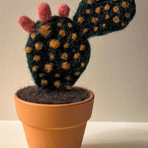 May include: A handmade felt cactus in a terracotta pot. The cactus is dark green with orange spots and pink accents. The pot is a light orange colour and filled with brown soil. A unique decorative item.
