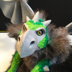 Könnte beinhalten: Eine graue und grüne Drachenpuppe mit einem pelzigen Körper und grünen Augen. Der Kopf des Drachen ist aus Ton und hat ein grünes und weißes Muster.