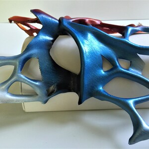 Pode incluir: Uma m&aacute;scara de couro azul e vermelho com um design delicado e intrincado. A m&aacute;scara &eacute; feita de couro fino e flex&iacute;vel e tem um brilho met&aacute;lico.