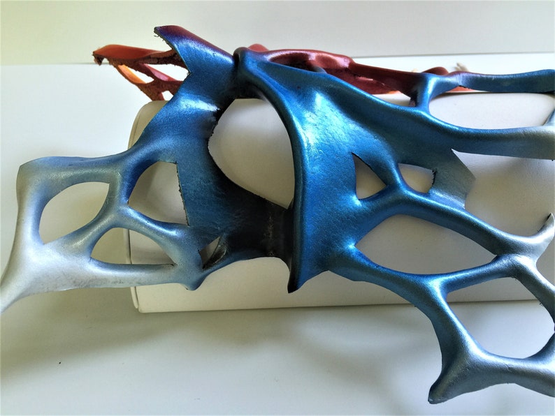 Pode incluir: Uma m&aacute;scara de couro azul e vermelho com um design delicado e intrincado. A m&aacute;scara &eacute; feita de couro fino e flex&iacute;vel e tem um brilho met&aacute;lico.