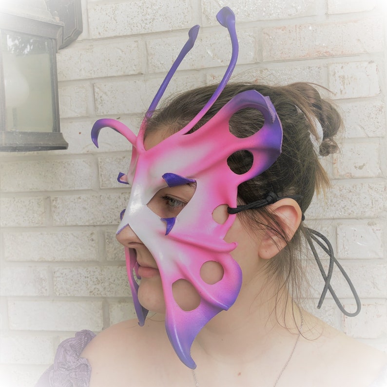 May include: A pink and purple butterfly-shaped masquerade mask with intricate details and a black elastic strap.