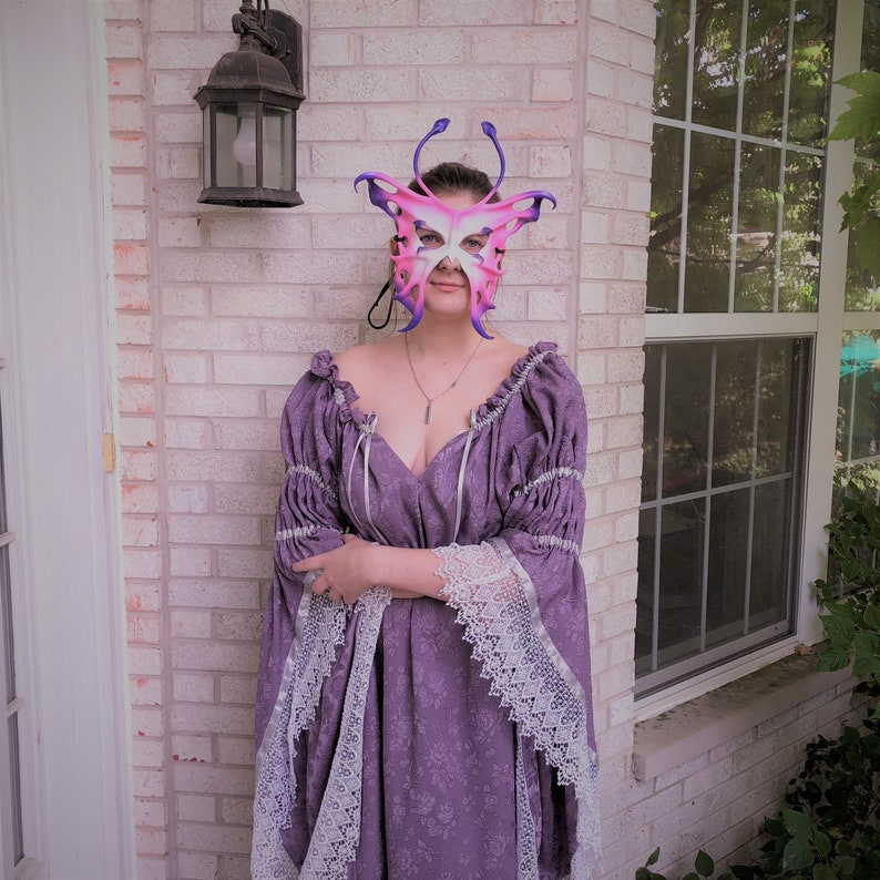 May include: A woman wearing a purple dress with white lace trim and a butterfly-shaped mask. The mask is pink and purple with a white design.