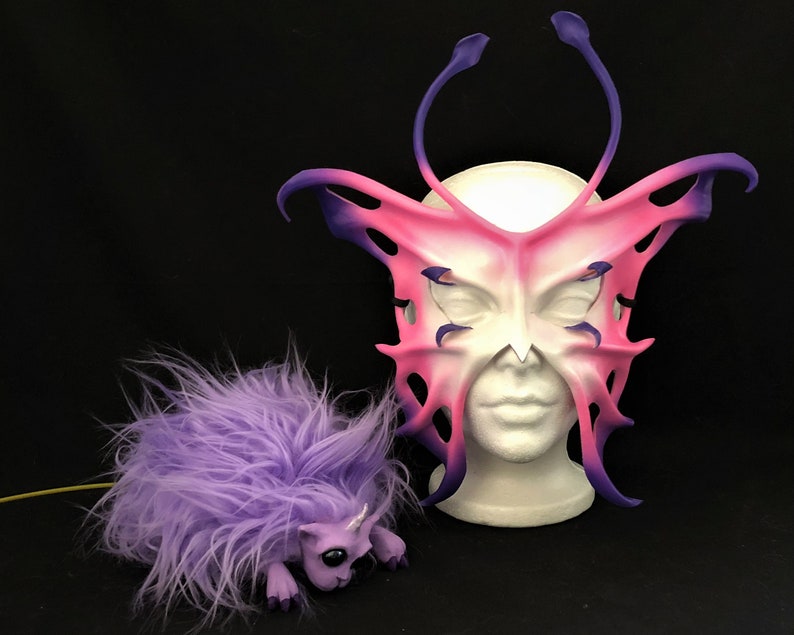 May include: A pink and purple butterfly mask with intricate details and a furry purple creature with a horn.