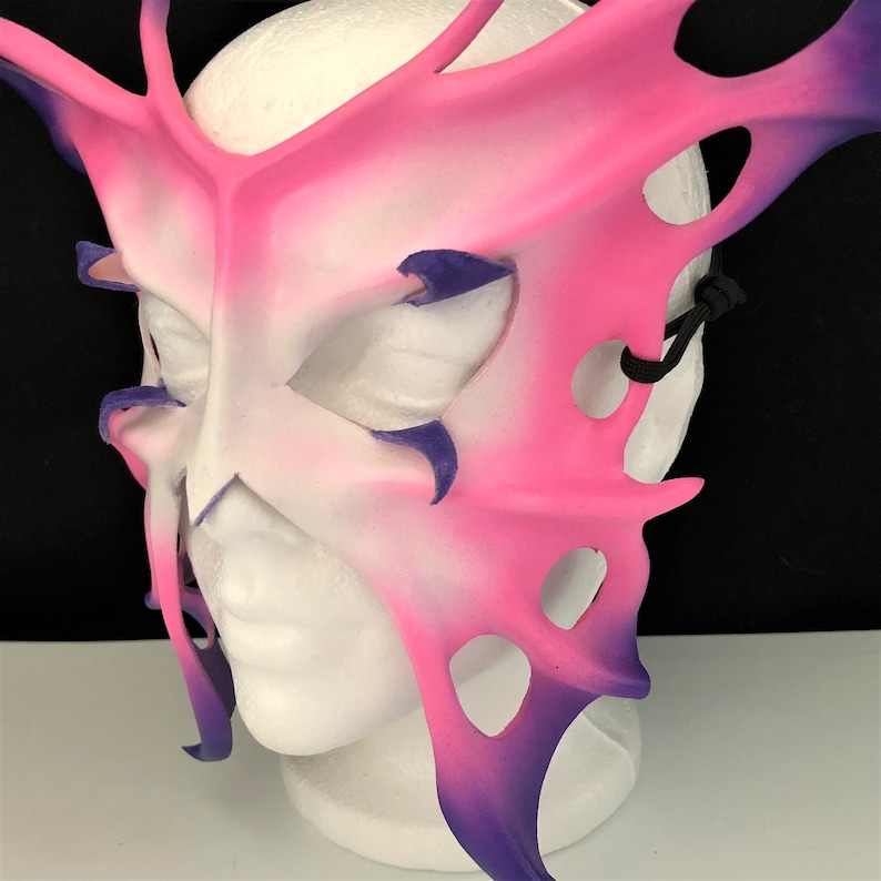 May include: A pink and purple butterfly-shaped masquerade mask with intricate details and cutouts. The mask is made of leather and has a black elastic strap.