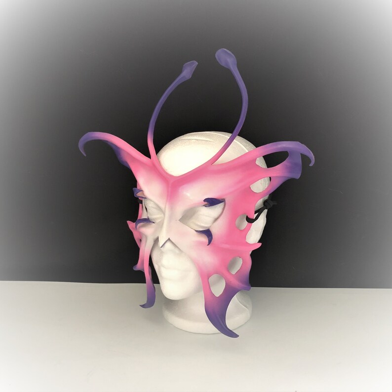May include: A pink and purple butterfly-shaped masquerade mask with intricate details and a delicate design. The mask is made of leather and has a three-dimensional effect.