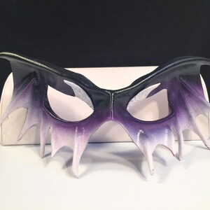 May include: A black and purple leather masquerade mask with a dramatic, pointed design. The mask has a gradient of color, fading from black to purple to white.