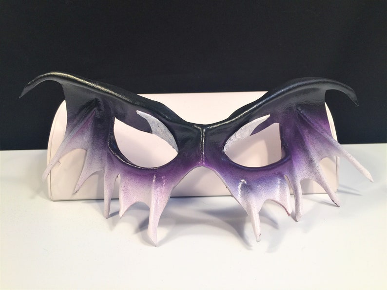 May include: A black and purple leather masquerade mask with a dramatic, pointed design. The mask has a gradient of color, fading from black to purple to white.