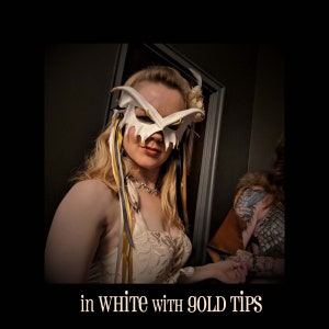 May include: A woman wearing a white masquerade mask with gold accents. The mask has a unique, angular design and is decorated with feathers.