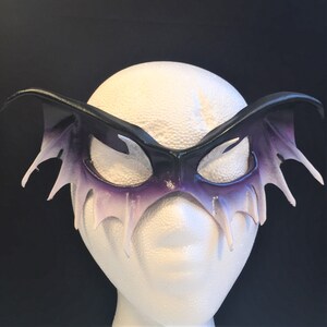 May include: A black leather masquerade mask with purple and white accents. The mask has a wing-like design with pointed edges.