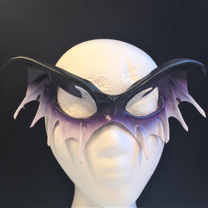 May include: A black leather masquerade mask with purple and white accents. The mask has a wing-like design with pointed edges.