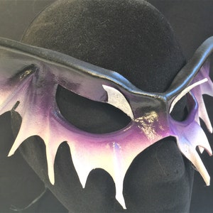 May include: A purple and white leather masquerade mask with a dramatic, spiky design. The mask is shaped like a bat with pointed wings and a long, pointed nose.