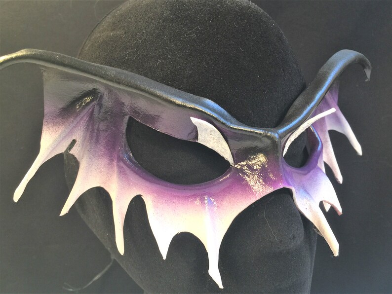 May include: A purple and white leather masquerade mask with a dramatic, spiky design. The mask is shaped like a bat with pointed wings and a long, pointed nose.