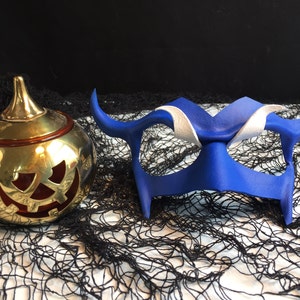 May include: A blue leather masquerade mask with white accents and horns, and a gold pumpkin-shaped candle holder with a red jack-o'-lantern face.