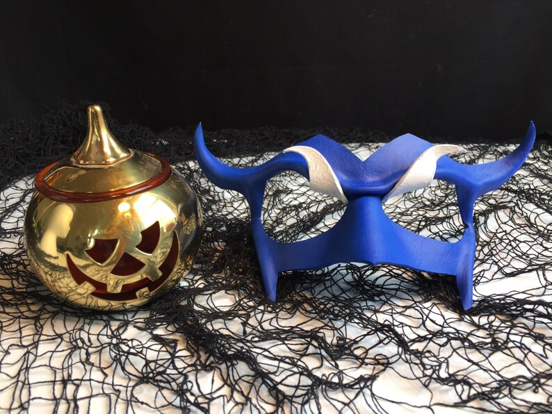May include: A blue leather masquerade mask with white accents and horns, and a gold pumpkin-shaped candle holder with a red jack-o'-lantern face.