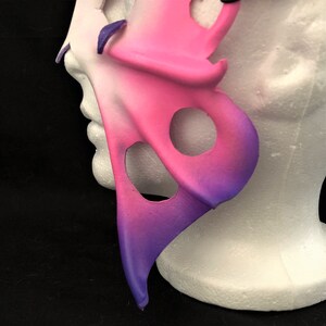 May include: A pink and purple leather masquerade mask with cutouts in the shape of circles. The mask is on a white polystyrene head.