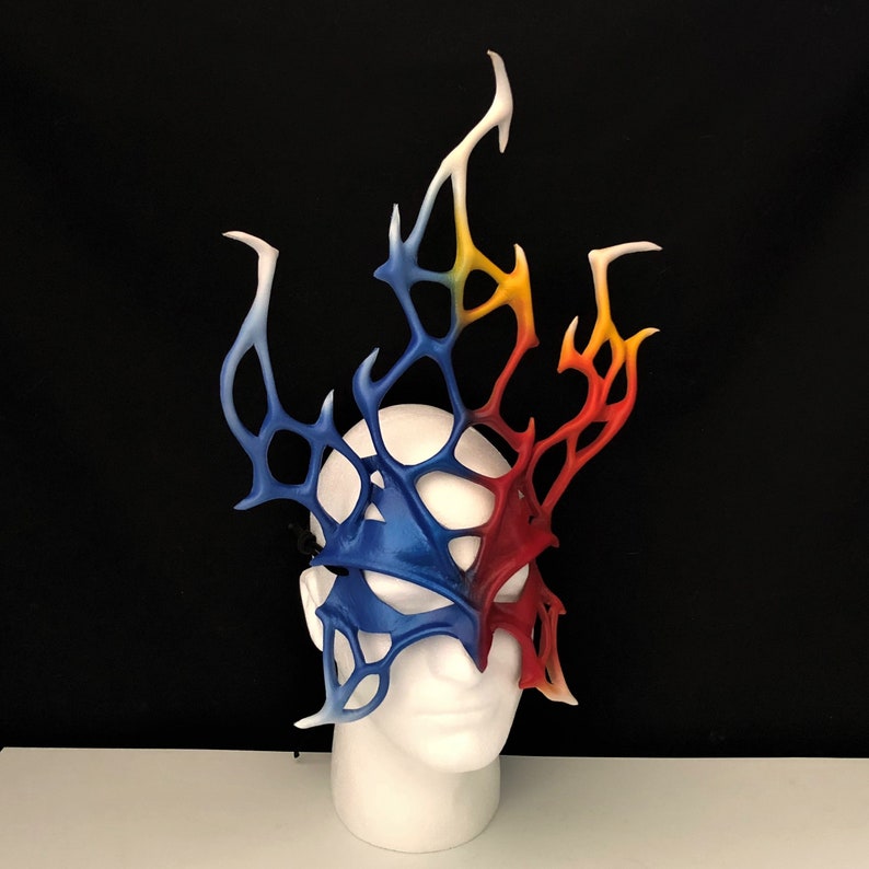 May include: A blue and red masquerade mask with a white, yellow, and orange flame design. The mask is made of a lightweight material and has a delicate, intricate design.