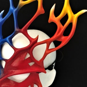 May include: A red, blue, and yellow leather mask with a white background. The mask is shaped like antlers and has a delicate, intricate design.