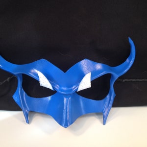 May include: A blue leather masquerade mask with white accents around the eyes. The mask has a pointed, angular design with horns on either side.