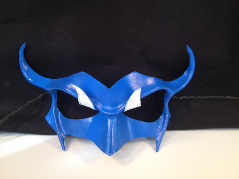 May include: A blue leather masquerade mask with white accents around the eyes. The mask has a pointed, angular design with horns on either side.