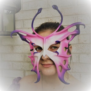 May include: A pink and purple butterfly-shaped masquerade mask with intricate details and a white centre. The mask has a black elastic band for wearing.