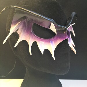 May include: A purple and white leather masquerade mask with a wing-like design. The mask is on a black mannequin head.