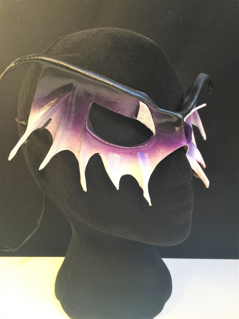 May include: A purple and white leather masquerade mask with a wing-like design. The mask is on a black mannequin head.