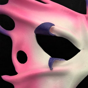 May include: A pink and white masquerade mask with purple accents. The mask has a large eye opening and a unique, abstract design.