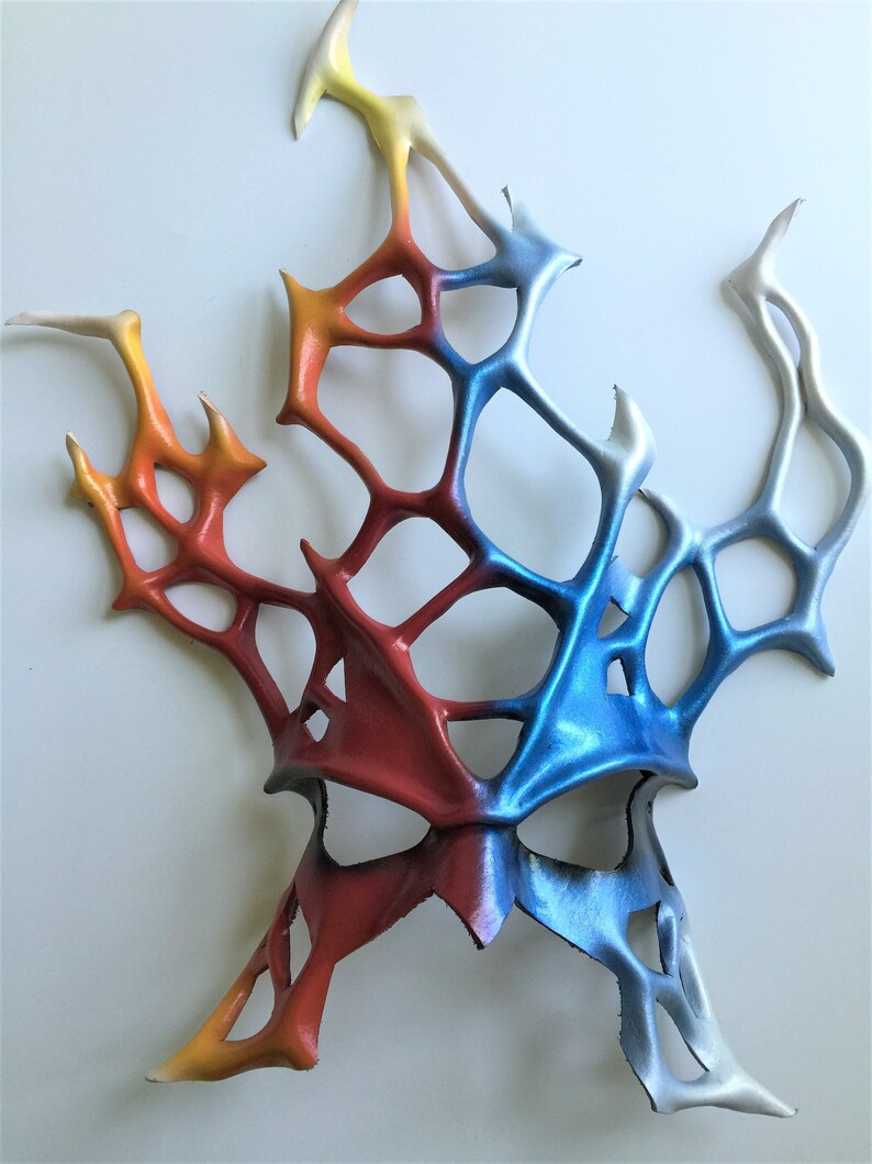 Pode incluir: Uma m&aacute;scara abstrata colorida feita de couro com um esquema de cores vermelho, laranja, amarelo, azul e branco. A m&aacute;scara tem um design geom&eacute;trico com um padr&atilde;o de teia de aranha.