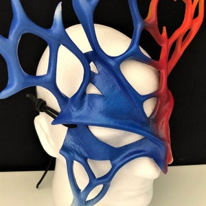 May include: A blue and orange masquerade mask with a detailed, intricate design. The mask is made of a lightweight material and has a black elastic strap.