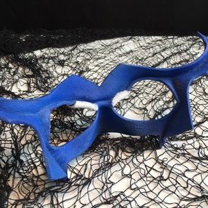 May include: A blue masquerade mask with a unique, abstract design. The mask has two large eye holes and a curved, pointed shape.
