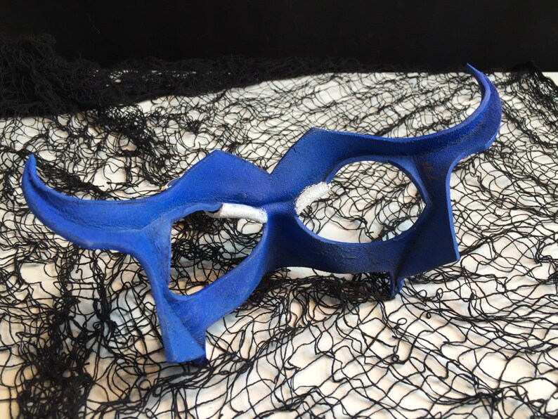 May include: A blue masquerade mask with a unique, abstract design. The mask has two large eye holes and a curved, pointed shape.