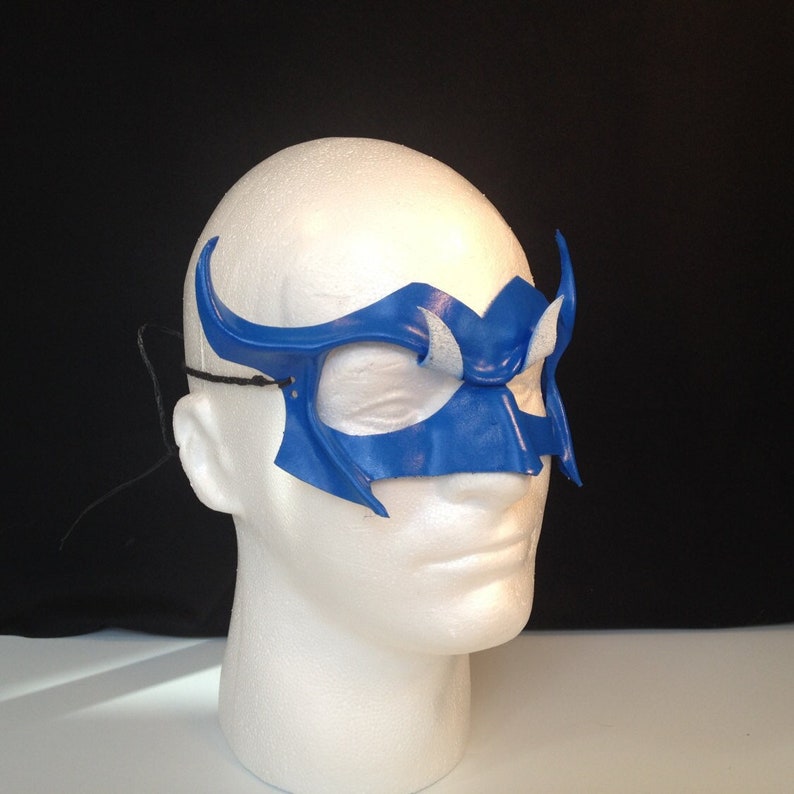 May include: A blue and white leather mask with a unique design. The mask has a pointed top and a curved bottom. It is worn on a white mannequin head.